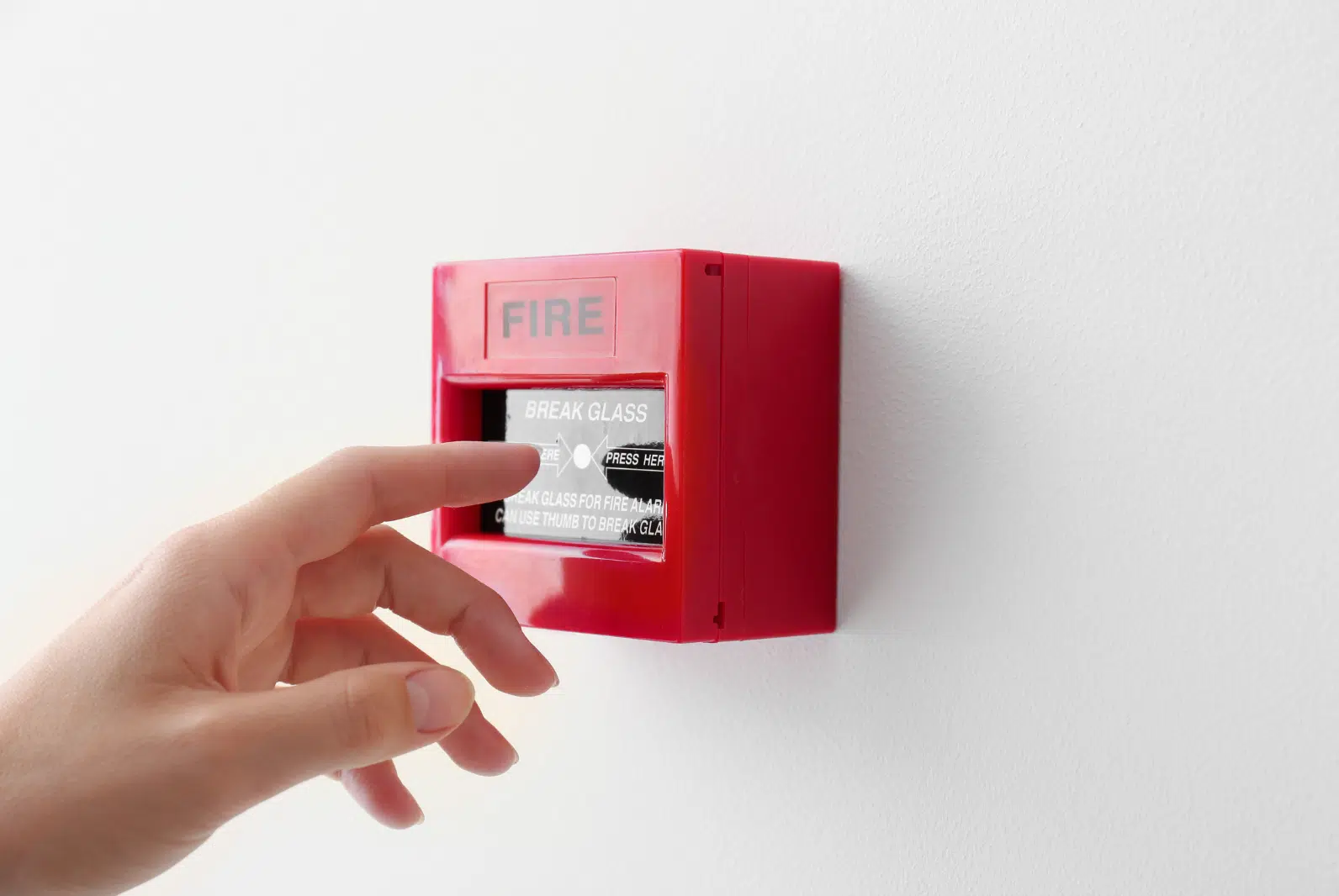 "Hand pressing a fire alarm break glass call point on a white wall for emergency fire safety activation."